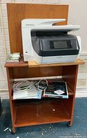 HP OfficeJet Pro 8720 printer on wooden stand with accessories on shelves below, including cords, paper, manuals, and ink cartridges.