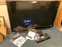 LG flat panel TV showing LG logo and 'No Signal' screen with remote controls, manuals, and BluRay player placed in front on the floor.