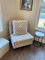 Modern armless chair with beige fabric featuring a white oval linked pattern, a large matching pillow, positioned near two windows and beside a round side table with a marble-like top.