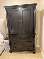 Front view of two-piece dark wood armoire featuring double doors with ring handles on the upper piece and three drawers with ring handles on the lower piece.