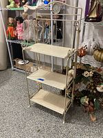 Front view of the aluminum bakers rack showing its three shelves and decorative scrollwork on the sides, located in a garage area.