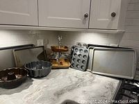 Photo of dark metal Bundt pan, silver metal Bundt pan, metal cupcake pan with 12 cups, two metal baking sheets, and the two-tiered wooden and pewter display stand on countertop