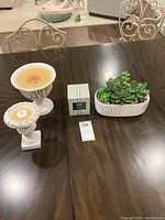 Two Nest Wellness candles in white distressed pedestal holders and one faux succulent plant on a dark wooden table.