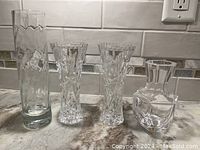 Four crystal vases of different shapes and cut glass patterns on marble counter under kitchen backsplash.