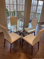Round glass top dining table set in a room with white chairs (not included). Visible wood floor and sliding glass doors.