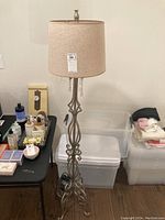 Full view of the lamp showing beige fabric shade, metal base with decorative scrolls, and crystal dangles hanging beneath the shade. One crystal is on the table.