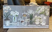 Framed painting with white floral arrangements on a shelf in jars and pots, neutral gray background, placed against a chair and brown carpet.