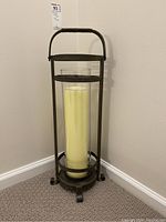 Front view of the wrought iron holder with the yellow hurricane candle inside placed on floor near wall.