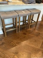 Photo showing three matching kitchen stools with wooden legs and distressed grey cushioned seats with studded trim.