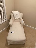 Front angled view of light colored chaise lounge with bolster pillow and President's Club blanket draped over backrest. Chair sits in corner of room on beige carpet near window with blinds.