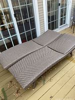 Two brown synthetic wicker outdoor chaise lounges placed adjacent on a porch deck, showing woven material and wheels on legs.