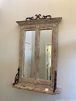 Front view of large rustic wooden wall mirror with two vertical mirror panels, distressed finish, and iron scroll brackets supporting a small shelf beneath the mirrors.
