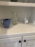 Three pitchers displayed on a white shelf with one blue ceramic floral pitcher, one clear textured glass pitcher, and one tall William Yeoward crystal pitcher with label.