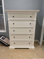Front view of a white tall dresser with five drawers showing decorative molding and black round knobs.