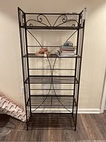 Front view of tall metal bakers rack bookshelf with five open shelves and decorative scrollwork on top and sides. Some books on middle shelf but described as contents not included.
