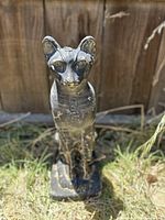 Full front view of the black carved cat statue showing facial features and body, placed outside on grass with wooden fence background.