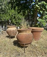 Three terra cotta pots outdoors on dry grass, two on metal stands, one sitting directly on ground