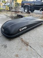 Black Inno brand rooftop cargo box on concrete, showing aerodynamic shape and branding.