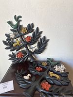 Two Mid-Century Modern tree of life metal wall hangings with colorful ceramic animal and flower decorations displayed against a white wall.