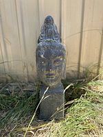 Front view of vintage large stone Buddha head statue standing in grass against a wall, showing facial details and base.