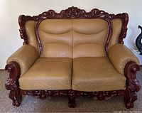Full front view of the vintage sofa showing tan leather upholstery and a richly carved dark wood frame with intricate floral and scroll details.