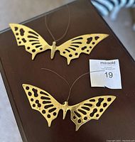 Two solid brass butterflies shown on brown surface, each with detailed cut-out wings and wire antennae.
