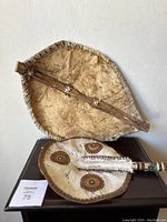 Two vintage African tribal shields displayed on a flat dark surface against a wall. One shield is plain hide with a wooden handle, the other has decorative circular patterns and a serrated handle.