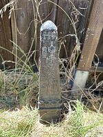 Front view of the obelisk showing carved Egyptian figure and hieroglyphs, set in grass outdoors with wooden fence background.