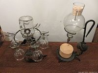 Two iron wine decanter stands side by side, one taller with a round glass decanter and wooden coasters at the base, the other shorter with an inverted decanter and six glasses hanging around it.