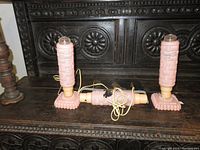 Three vintage pink glass lamps arranged on dark carved wood surface. Two table lamps stand upright; one lays flat between them with cords attached to two. Each lamp has basket weave pattern on the body and floral scalloped base.