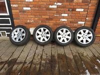 Four silver BMW factory alloy wheels lined up against a brick wall on wooden floor. All four wheels have used tires mounted.