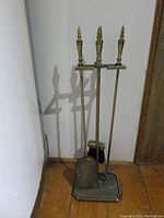 Full view of the brass fireplace tool set including the stand, poker, brush, and scoop placed on wooden floor near wall.