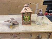 Photo of copper candle holder, wooden bird figurine, and glass dispenser on wooden surface with cardboard background.