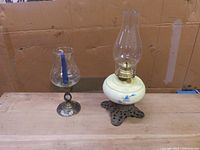 Photo showing ceramic and brass oil lamp with white hand-painted font with blue flowers, clear glass chimney on an ornate brass base, and brass-based candle holder with glass hurricane enclosing a blue candle.