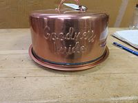 Side and angled view of shiny copper cake cover with embossed "Goodness Inside" text, showing the smooth polished surface and round handle on top.