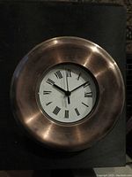 Copper coated round clock with visible corrosion and tarnish on the frame. White clock face with black Roman numerals and three black clock hands.