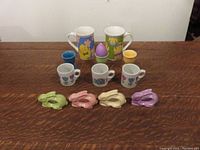 Photo shows 2 large mugs with Easter designs, 3 small mini mugs with floral and Easter motifs, 3 egg cups including 1 purple egg candle holder, and 4 pastel-colored ceramic bunny napkin holders.