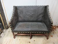 Front view of two-seater settee with embossed leather upholstery and brass nailhead trim, showing intricate leatherwork and wooden legs.