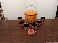 Photo showing six matching orange ceramic mugs with black interiors and black jack-o'-lantern face designs, one glass pumpkin-scented candle with smiling pumpkin face, and one large orange metal Halloween loot bucket with lid and metal handle, all arranged on a wooden table.