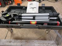 Top view inside the storage case showing the adjustable tripod stand, Jobmate level base with bubble level, circular level, and instruction manual laid out inside.