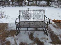 Front view of metal rocking bench showing decorative circular designs on backrest, curved armrests, and metal slat seat. Snow and concrete floor visible.
