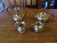 Four-piece silver plated tea set displayed on wooden table under warm light: coffee pot, teapot, creamer, sugar bowl