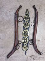 Photo showing two hardwood horse traces with iron hardware and a black leather belt decorated with five solid brass medallions and buckles laid on concrete floor.
