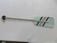Photo of paddle front showing light green color with black diagonal stripes, wooden handle and hanging loop.