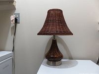 Photo of lamp against wall, showing full lamp with glass base and wicker shade