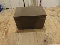 Closed wooden rectangular painted storage box on wooden floor. Brown color with no visible markings on top or sides.