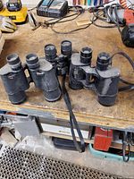 Three black binoculars on a workbench with tools around. Two are paired closely together, showing their front lenses.