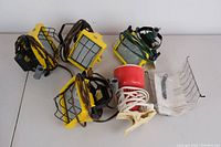 Photo of four yellow halogen work lights with black protective metal cages and cables, alongside one red clamp lamp with white power cord and clamp base.