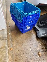 Stack of 5 blue plastic storage bins with hinged lids seen from side angle on concrete floor.