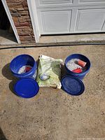 Overview photo of two blue buckets with blue lids, two red plastic cups inside buckets, and a biodegradable bag of ice melt between them on a concrete floor outside a garage door.